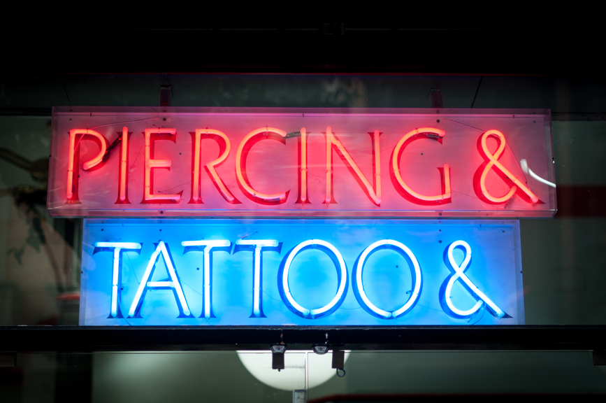 Two luminous signboards on building at night. Text with Tattoo in blue colour. Red sign with Piercing written on it. Modern dark building as background. Popular activity for young people.